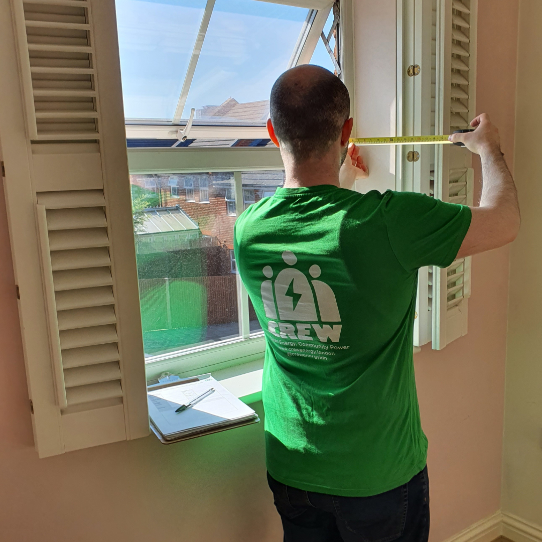 A man wearing a green tshirt measures a window with a yellow tape measure. On the window sill is a clipboard which holds a notebook full of measurements.